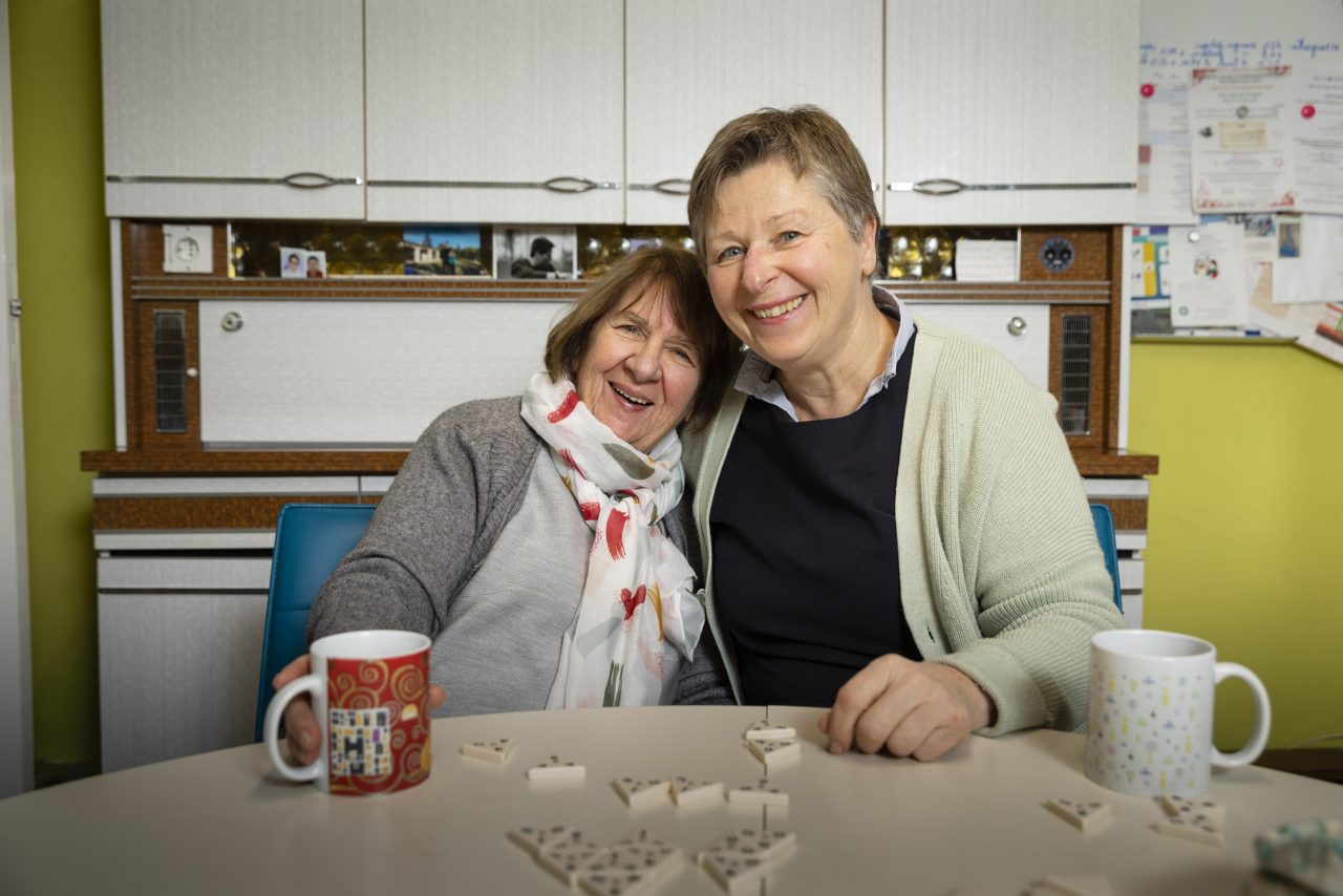 Une mère atteinte de la maladie d'alzheimer avec sa fille aidante à ses côtés sont assises à la table de la cuisine devant un jeu de société à l'heure du thé, elles regardent l'objectif en souriant