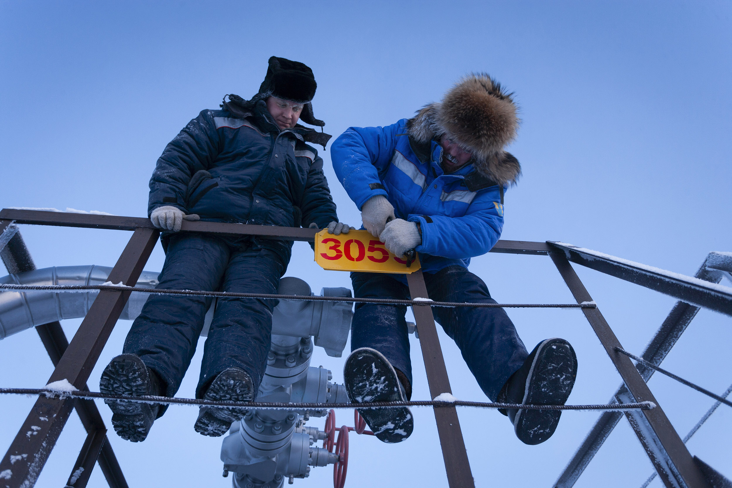 Photographie professionnelle en contre-plongée de 2 hommes au travail sur un stockage de gaz en Sibérie l'hiver par moins 43 degrés sous un ciel bleu