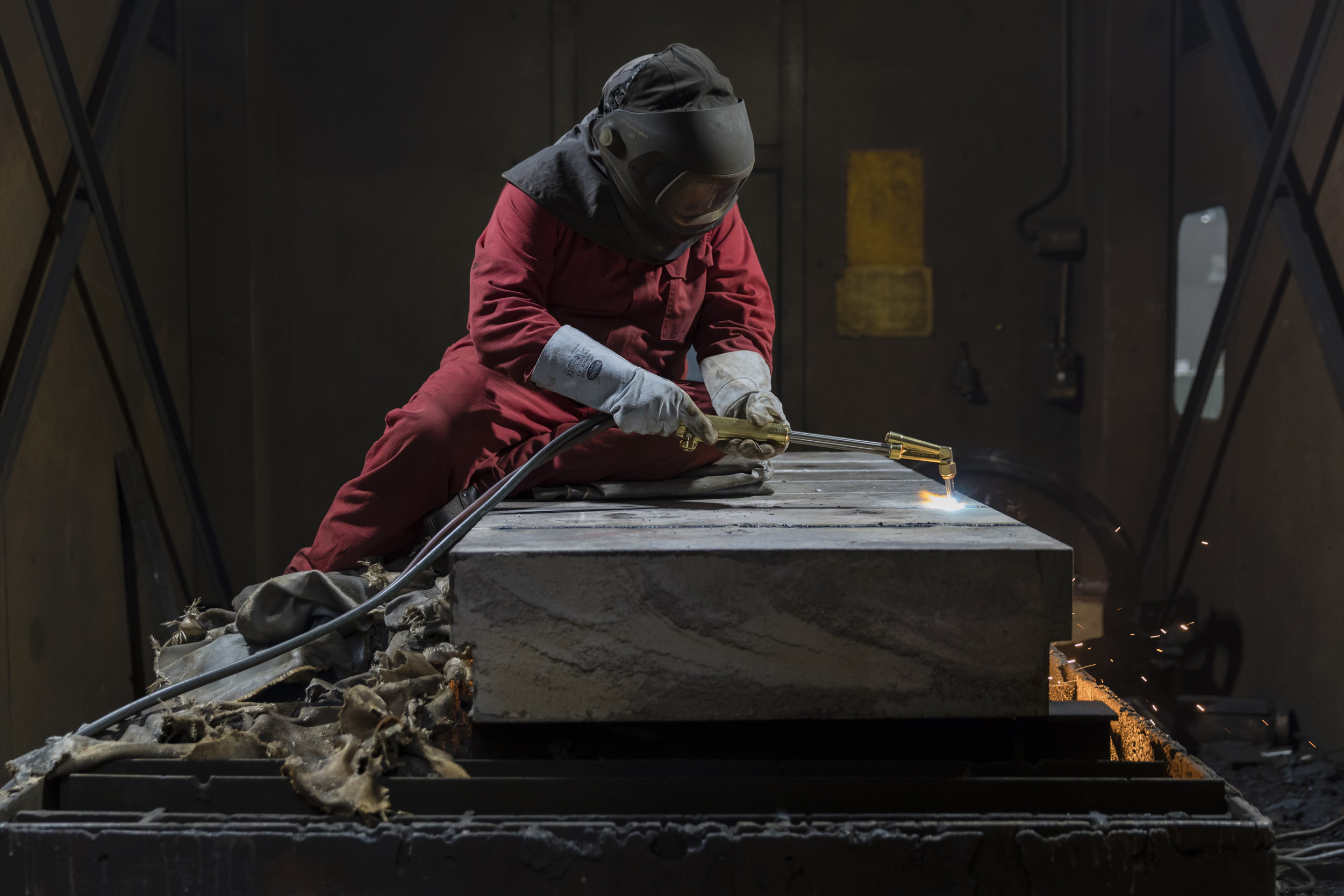 Photographie industrielle d'un oxycoupeur en tenue de travail qui découpe au chalumeau une plaque d'acier très épaisse sur laquelle il est installé