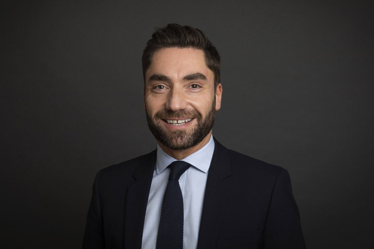 Portrait corporate cadré en plan poitrine d'un homme souriant en studio sur fond gris foncé. Il porte une chemise bleu ciel, une cravate et une veste.