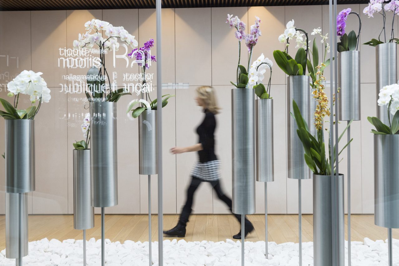 Femme en mouvement dans un patio derrière une série de vases design en inox contenant des orchidées colorées
