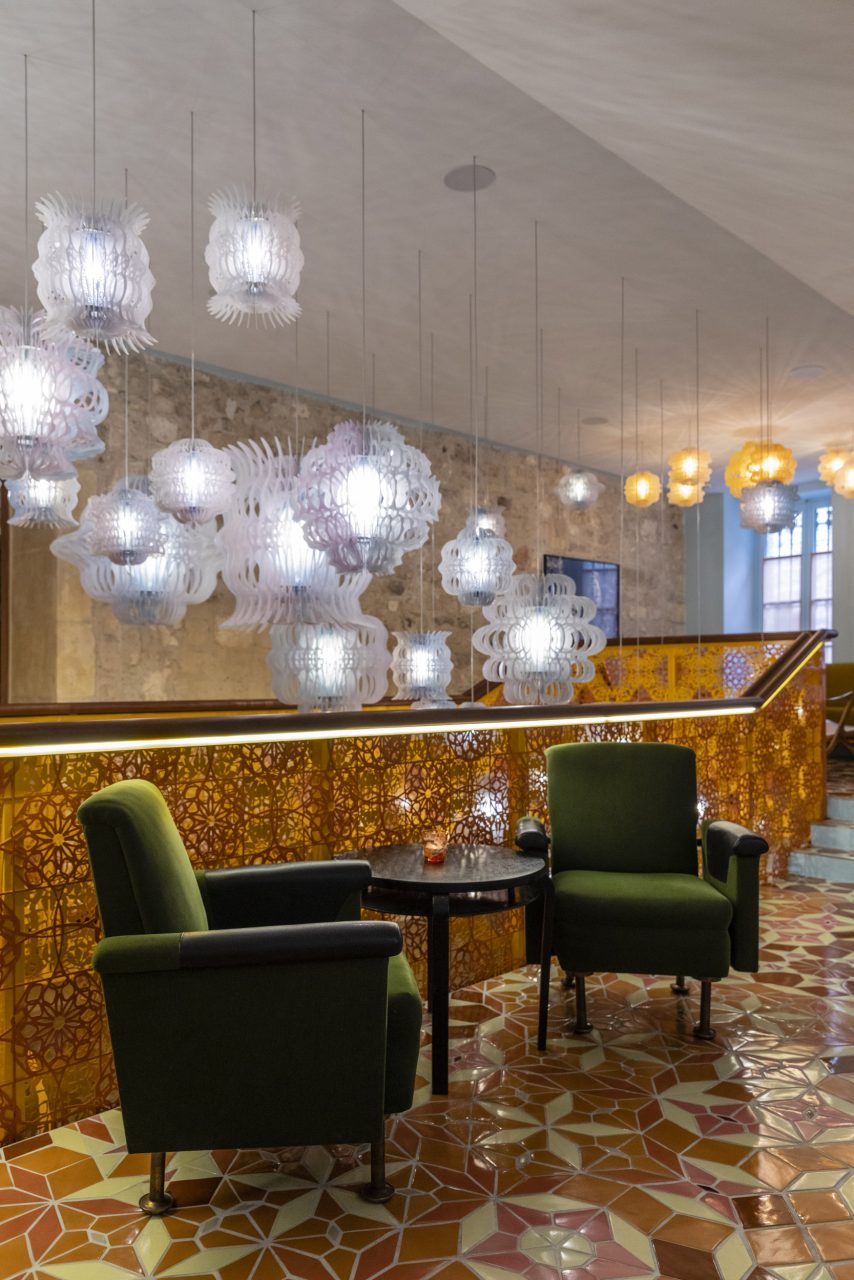 2 fauteuils de couleur verte et une table basse dans un hôtel de luxe à la décoration moderne, le carrelage au sol est jaune et orange, des luminaires en verre sont suspendus au plafond à l'arrière plan